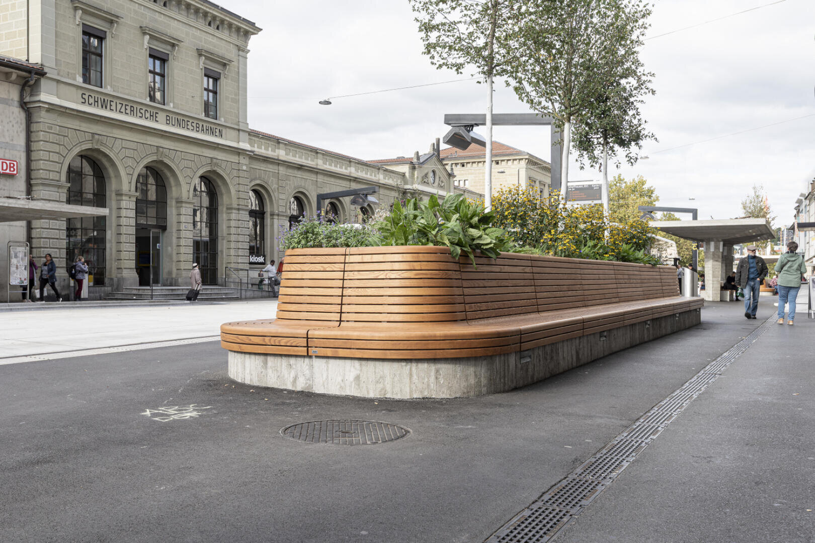 A long, curved wooden bench with built-in planters and green plants stands on a sidewalk near the Swiss Federal Railways station. People are walking nearby and the stone façade of the station can be seen in the background.