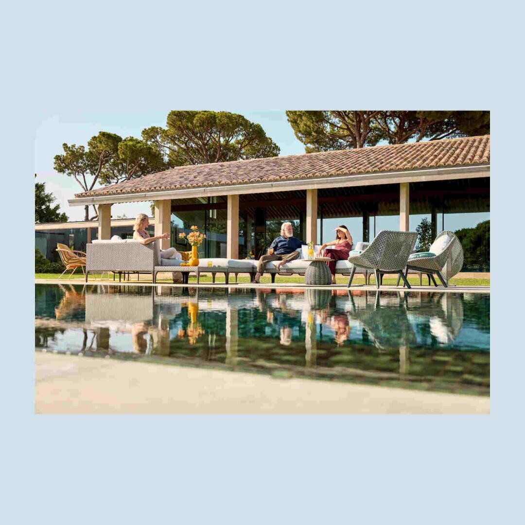 Drei Personen entspannen sich auf modernen Gartenmöbeln an einem Pool, unterhalten sich und genießen Getränke, während sich im Hintergrund ein großes Haus und Bäume im Wasser des Pools spiegeln.