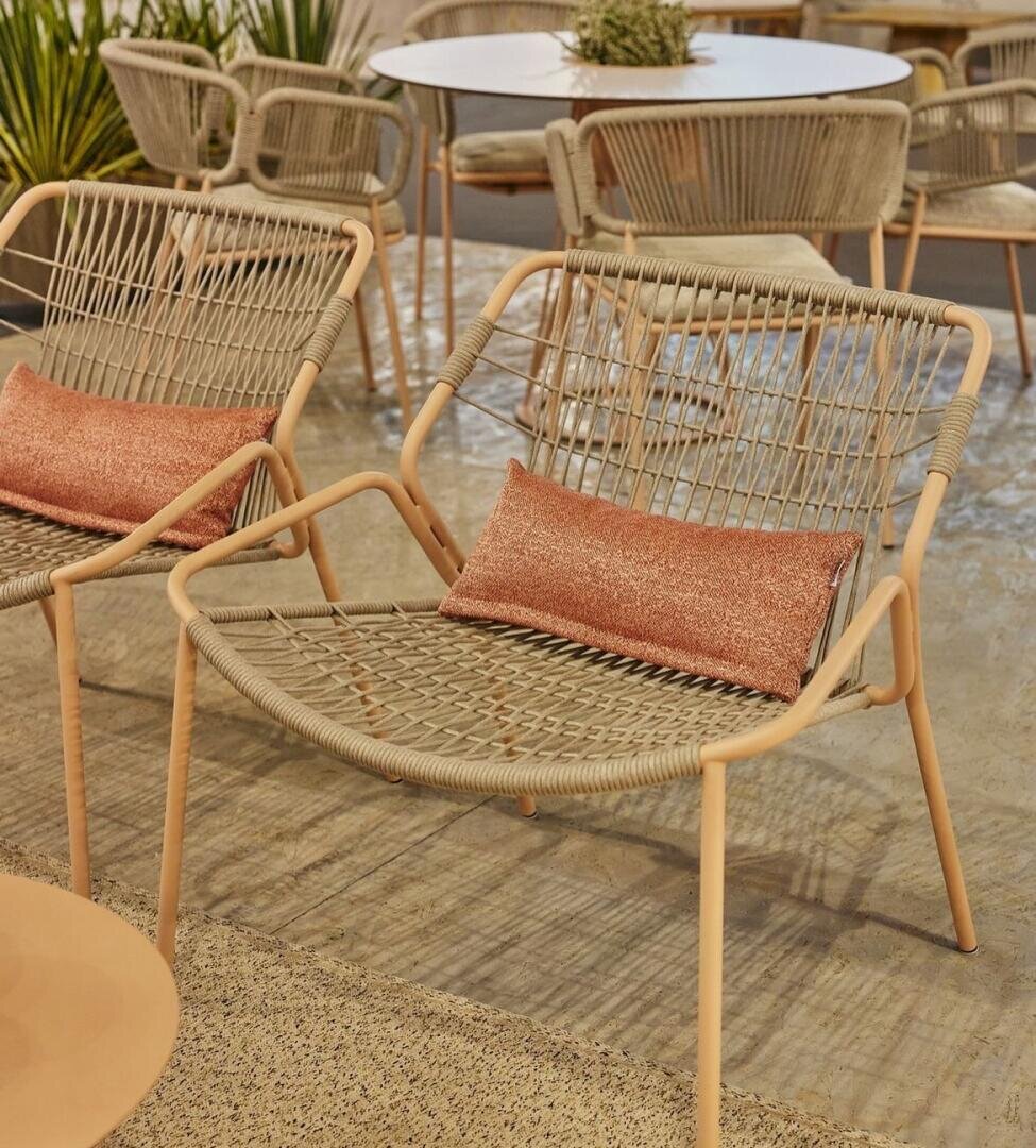 Zwei moderne geflochtene Loungesessel mit hellbraunem Gestell und rosafarbenen rechteckigen Kissen stehen in einem Innenraum auf einem strukturierten Boden. Im Hintergrund sind ein runder Tisch mit Stühlen und Grünpflanzen zu sehen.