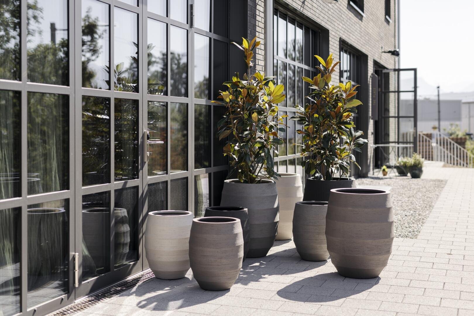 Vor einem modernen Gebäude mit großen Glasfenstern und Backsteinmauern stehen an einem sonnigen Tag große Keramikkübel, einige leer, andere mit grünen Pflanzen.