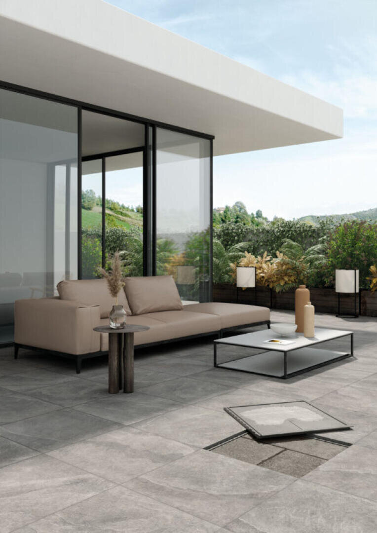 Moderne Außenterrasse mit einem beigen Sektionssofa, einem niedrigen Couchtisch und einem Beistelltisch auf grauem Fliesenboden; einige Fliesen sind angehoben, um die darunter liegenden Schichten zu zeigen. Im Hintergrund sind Glastüren und üppiges Grün zu sehen.
