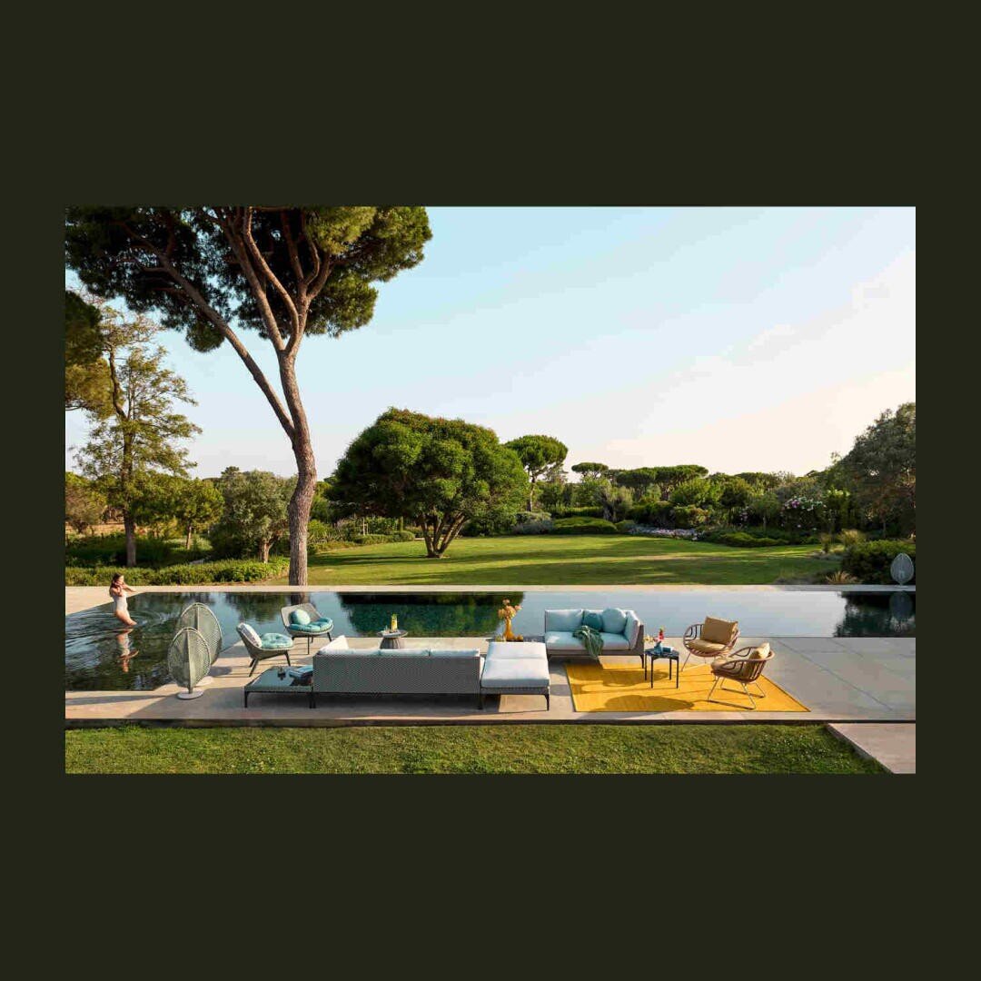 Ein moderner Lounge-Bereich im Freien mit Sofas, Stühlen und einem gelben Teppich befindet sich neben einem spiegelnden Pool. Üppig grüne Bäume und Gras füllen den Hintergrund unter einem klaren Himmel.