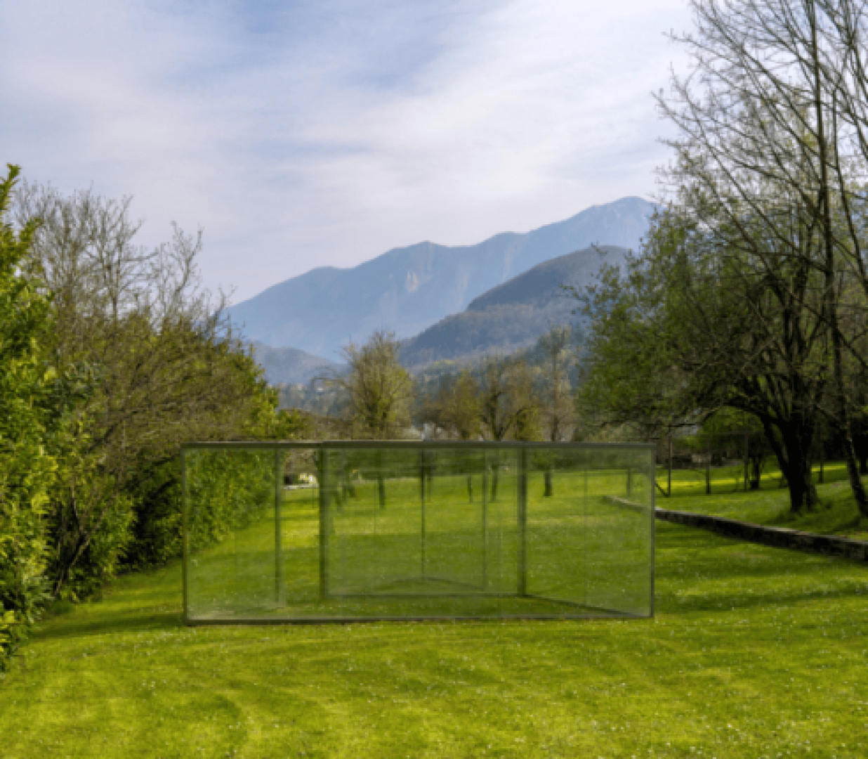 Ein durchsichtiger Glaskasten steht auf einer grünen Wiese, umgeben von Bäumen, mit Bergen und einem bewölkten Himmel im Hintergrund.