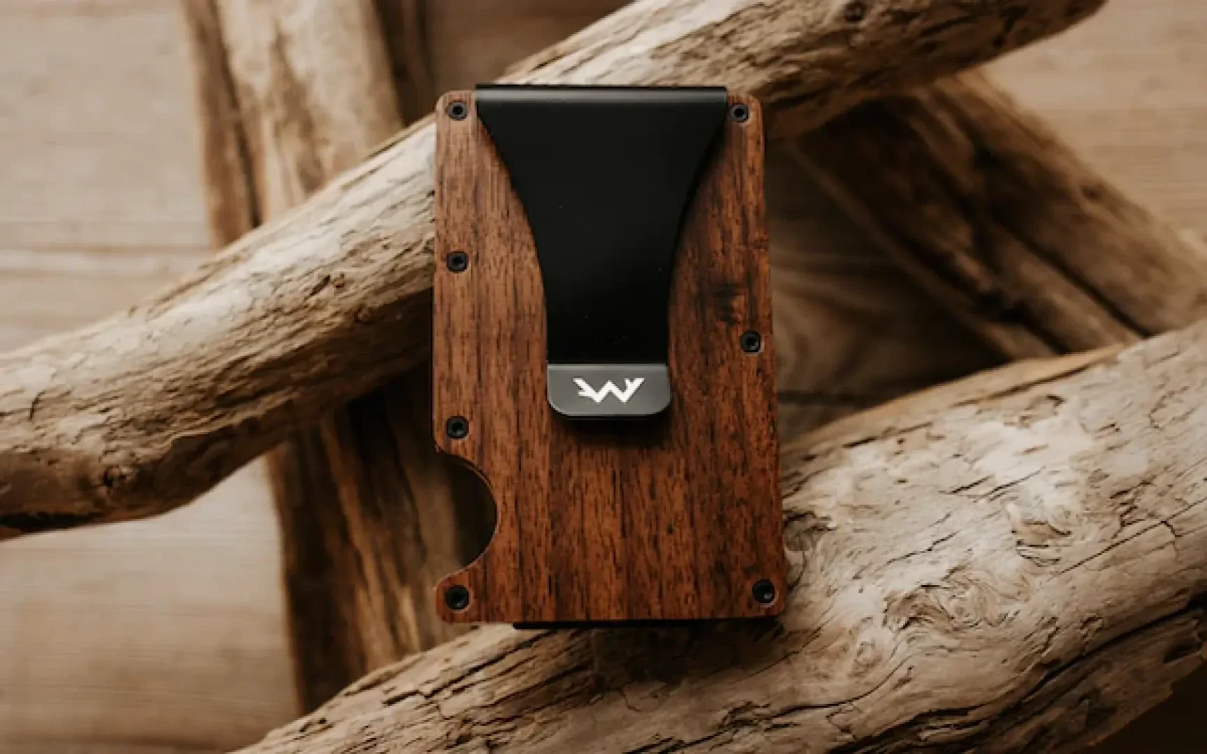 A slim, wooden wallet with a black metal clip and a white logo, resting on light-colored, textured driftwood pieces.