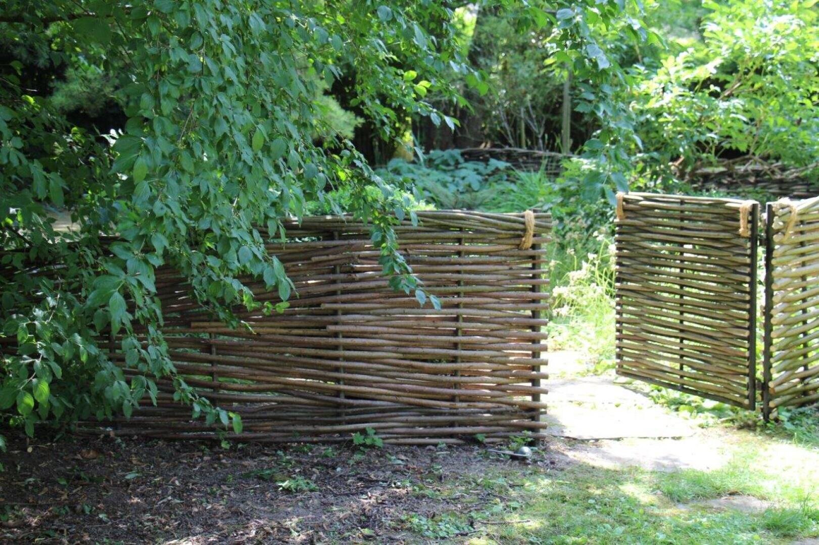 A woven wooden fence with an open gate stands in a lush, green garden, partially shaded by overhanging branches and surrounded by dense foliage. The sunlight shines through the leaves onto the grass and the path.