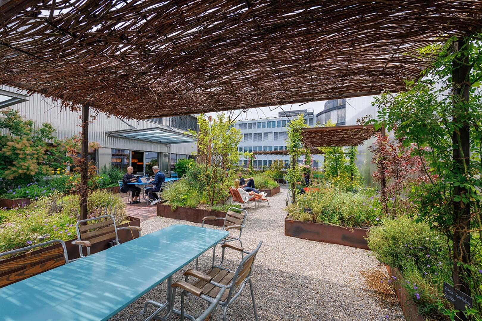 Sitzbereich im Freien mit Holztischen und -stühlen, umgeben von üppigem Grün, Pflanzen in Hochbeeten und einer geflochtenen Pergola über dem Dach. Im Hintergrund sitzen Menschen und unterhalten sich an einem sonnigen Tag.