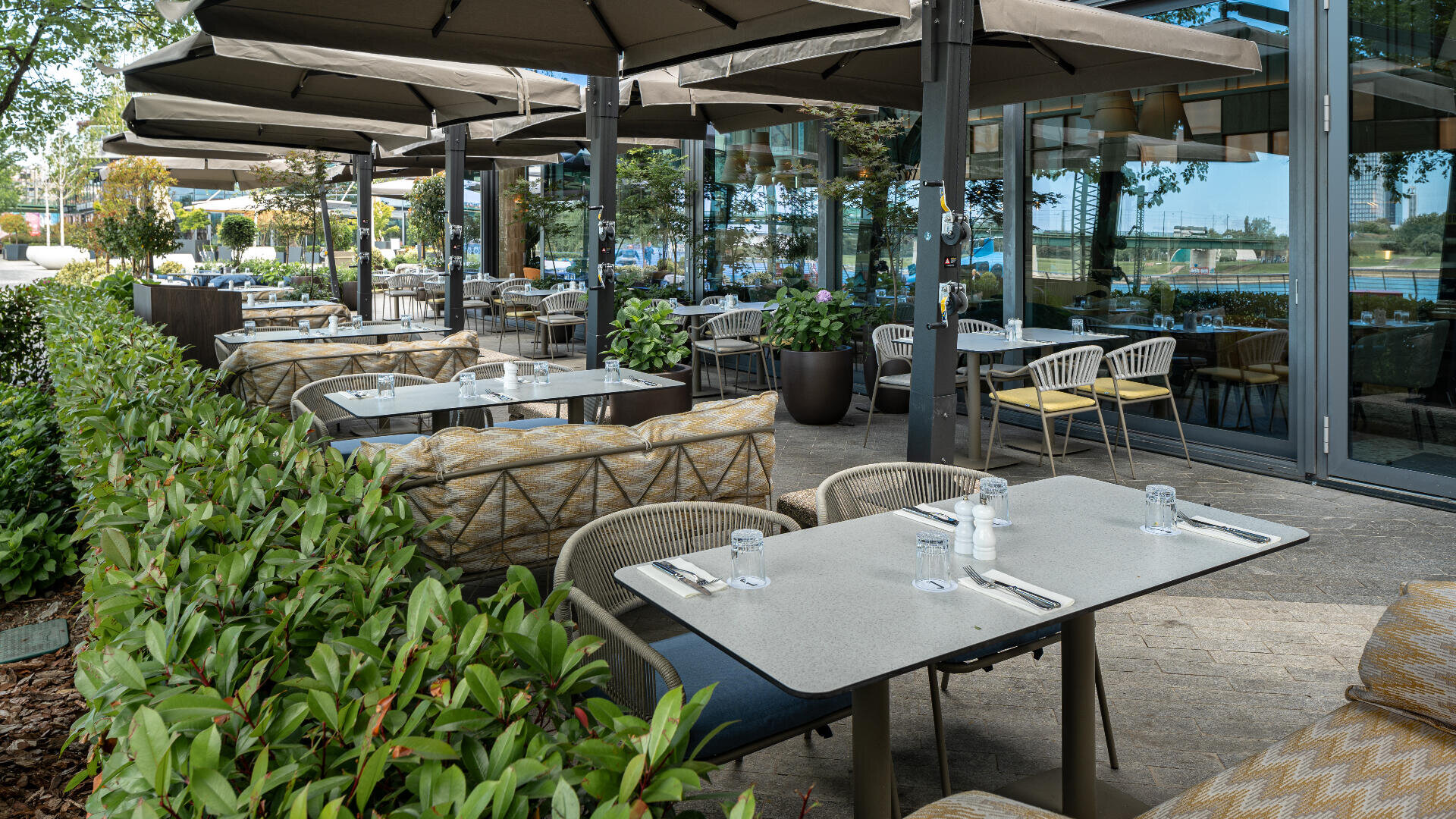 Das Restaurant verfügt über einen Außenbereich mit modernen Tischen und Stühlen, Gedecken und großen Sonnenschirmen. Der Bereich ist von Grün umgeben und liegt neben einem Glasgebäude, in dem sich Bäume und Himmel spiegeln.