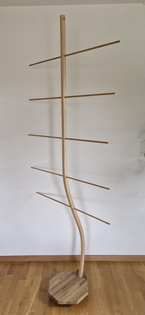 Ein hölzerner Kleiderständer mit einer gedrehten zentralen Stange, sechs horizontalen Ästen auf jeder Seite und einem achteckigen Sockel, der auf einem Holzboden vor einer weißen Wand steht.