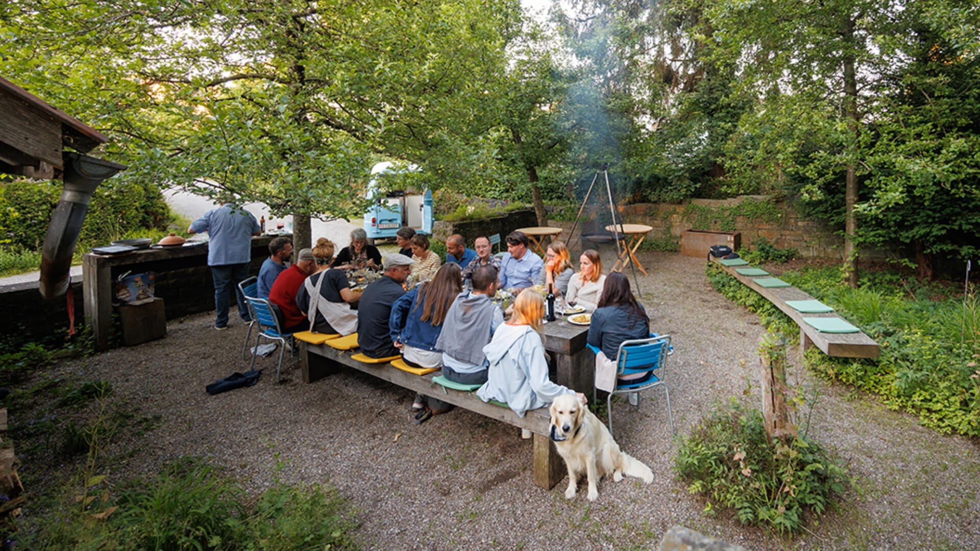 Eine Gruppe von Menschen sitzt an einem langen Tisch im Freien und isst gemeinsam in einem Garten, umgeben von Bäumen und Grünpflanzen. Ein Hund sitzt in der Nähe des Tisches, und im Hintergrund steigt Rauch von einem Grill auf.