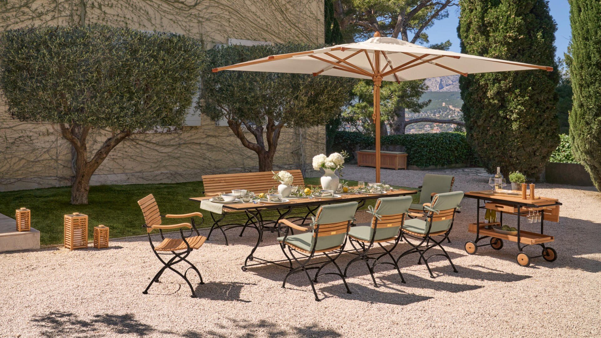 Dining area on the terrace with a wooden and metal table, six chairs and a bench under a large beige parasol. The table is set for a meal, surrounded by greenery and trees on a gravel surface.