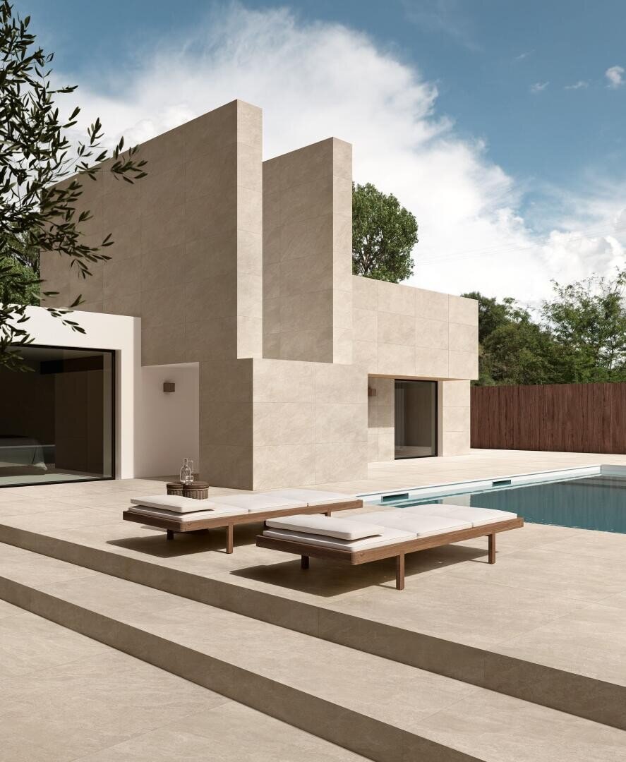 Modern, minimalist house with beige stone façade, large geometric shapes and high windows; two upholstered wooden armchairs stand next to a rectangular swimming pool on a spacious terrace under a partly cloudy sky.