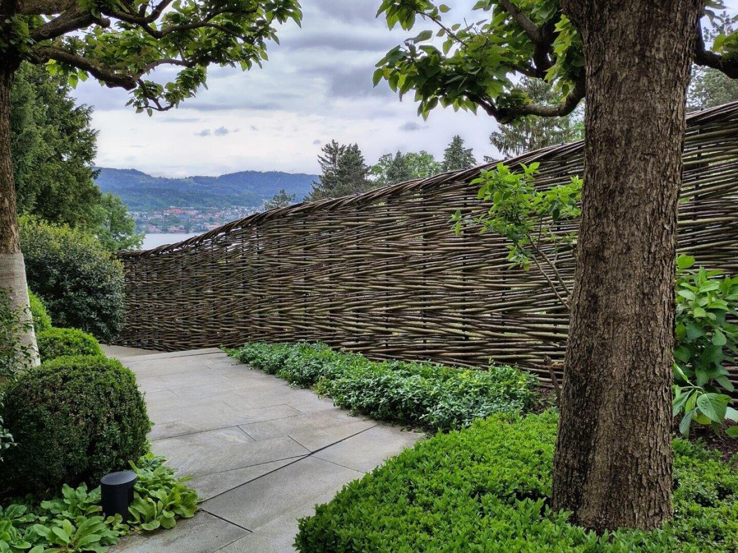 A stone path, lined with neatly trimmed bushes and trees, meanders past a high, woven wooden fence, while hills and a cloudy sky can be seen in the background.