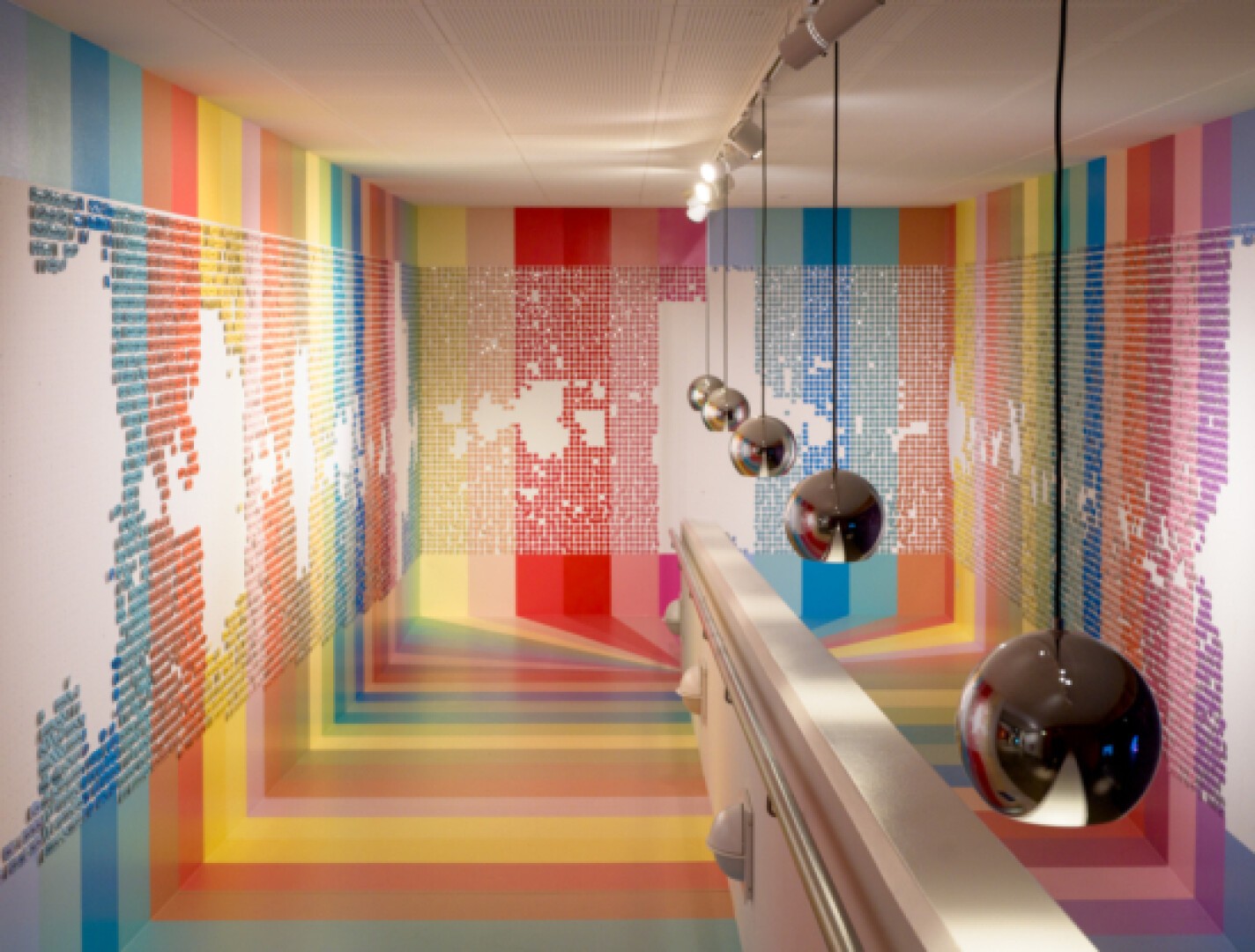 Ein farbenfroher, geometrischer Raum zeichnet sich durch Regenbogenstreifen an Wänden, Decke und Boden aus. Von der Decke hängen verspiegelte Kugeln, die die lebendige Umgebung reflektieren. Im Vordergrund verläuft ein weißes Geländer.