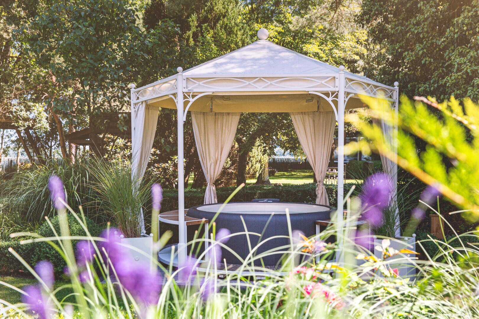 A white pavilion with open curtains stands in a garden surrounded by greenery, tall grass, and blooming purple flowers, creating a peaceful outdoor atmosphere.