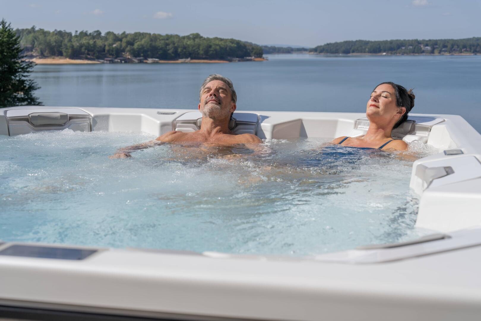 Ein Mann und eine Frau entspannen sich in einem Whirlpool mit Blick auf einen malerischen See mit Bäumen im Hintergrund unter einem klaren Himmel. Beide haben ihre Augen geschlossen und wirken friedlich.