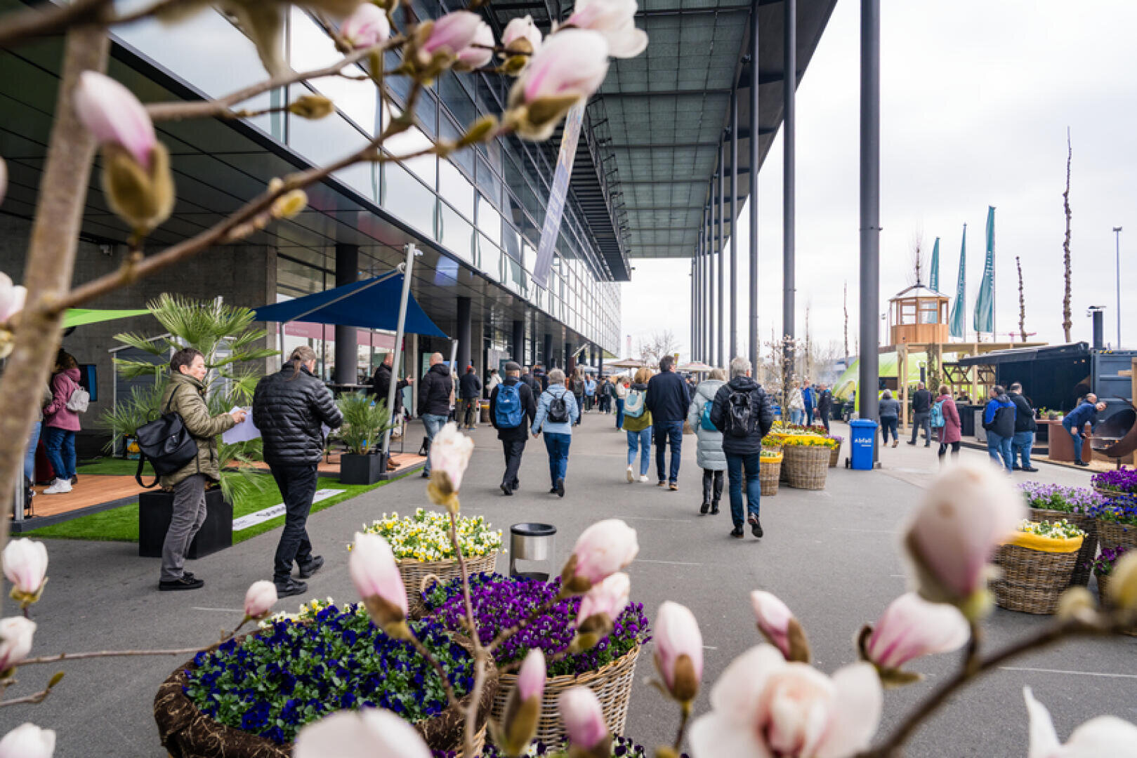 An einem bewölkten Tag gehen Menschen vor einem modernen Gebäude spazieren, umgeben von Pflanzkübeln mit blühenden Blumen und Magnolienzweigen im Vordergrund. Einige Besucher machen Fotos und andere erkunden die Gegend.