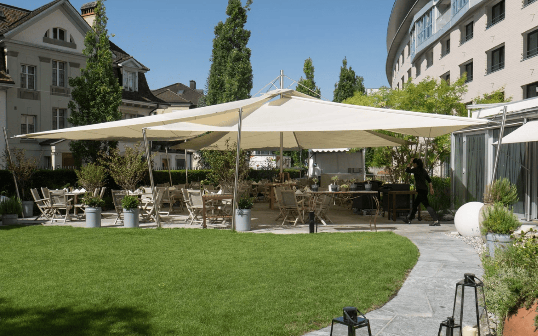 An outdoor dining area with wooden tables and chairs is shaded by large cream-colored canopies. The space is surrounded by greenery and buildings, with a person walking near the seating area.