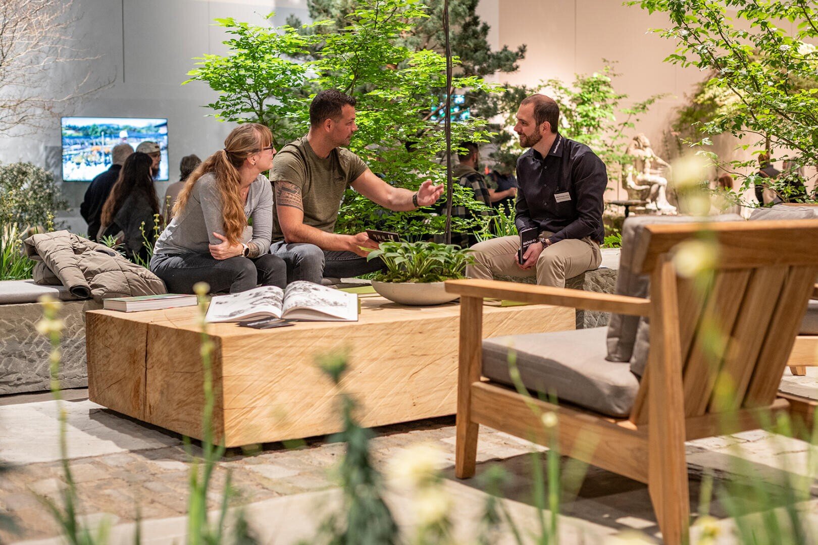 Drei Personen sitzen um einen Holztisch im Freien mit Pflanzen und einem aufgeschlagenen Buch, umgeben von üppigem Grün und Holzmöbeln in einer modernen Gartenanlage und unterhalten sich.
