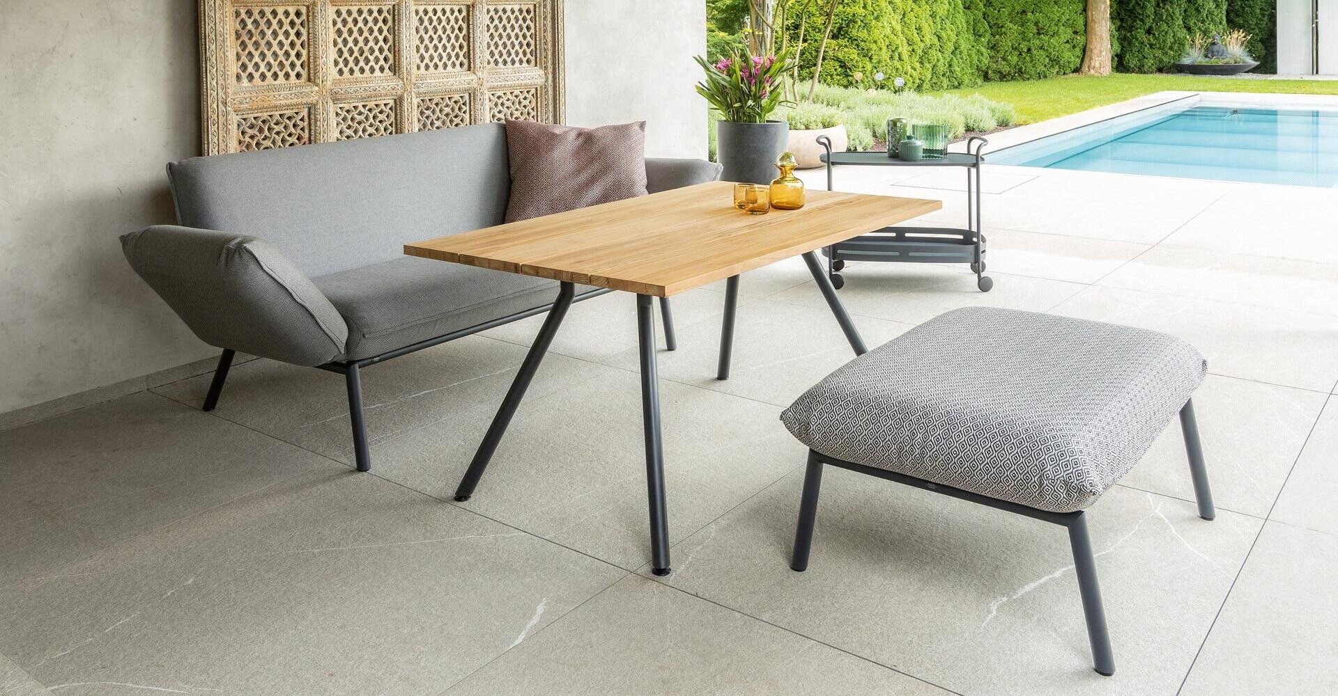 Moderne Außenterrasse mit einem grauen Sofa, einer passenden Ottomane, einem Holztisch, einem Servierwagen und Topfpflanzen neben einem Swimmingpool an einem sonnigen Tag.