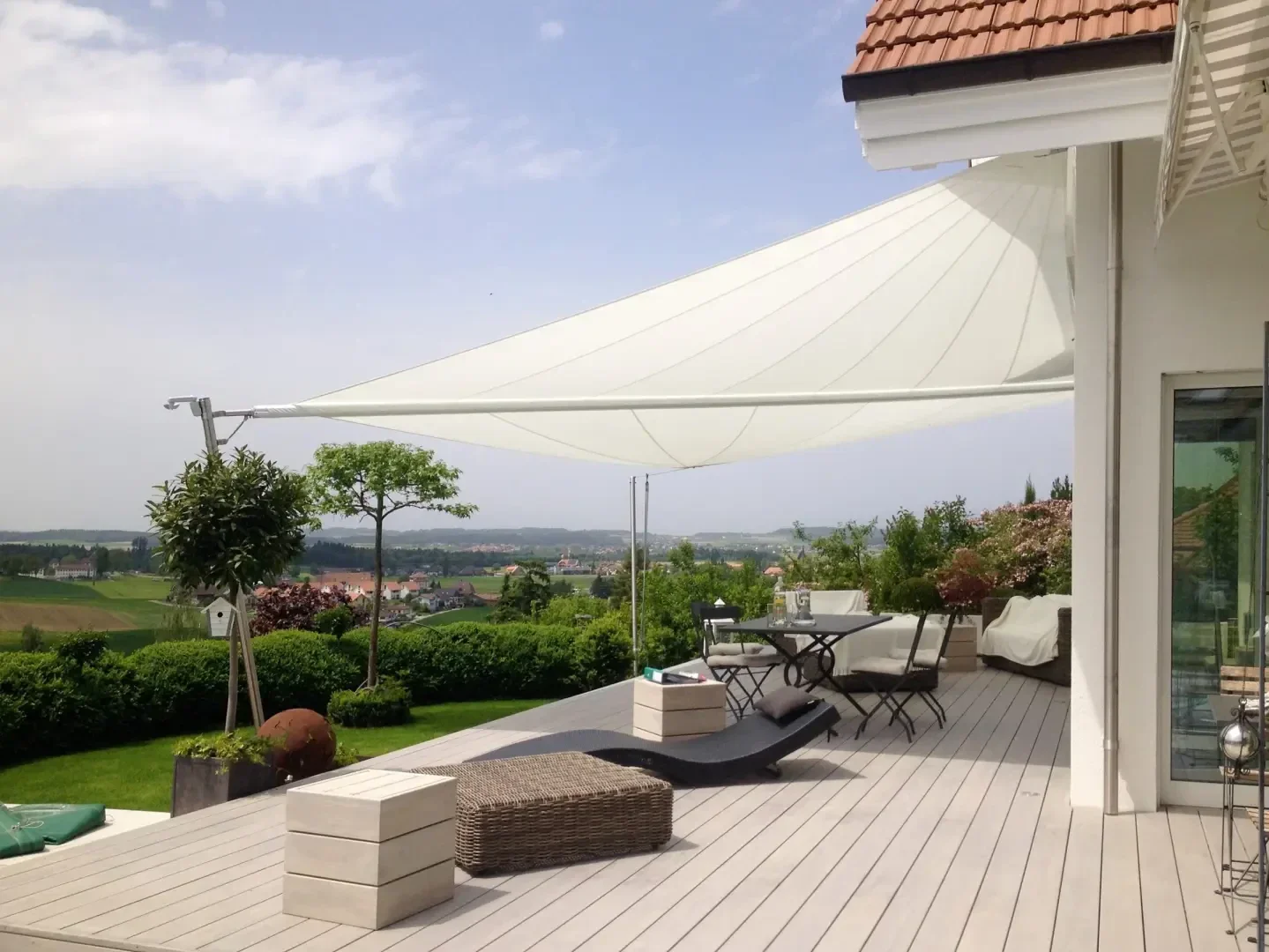 Moderne Terrasse mit hellem Holzbelag, stilvollen Außenmöbeln und einem großen weißen Sonnenschirmdach. Der Bereich überblickt eine malerische Landschaft mit Feldern und fernen Hügeln unter einem teilweise bewölkten Himmel.