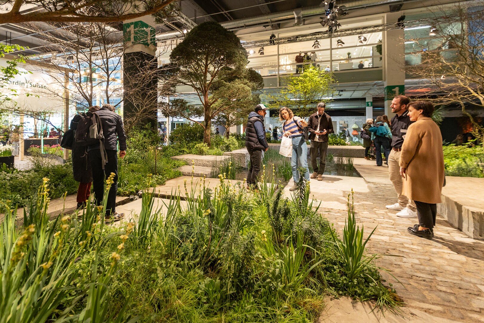 Menschen spazieren durch einen Indoor-Garten mit Pfaden, Grünpflanzen und Bäumen. Einige beobachten die Pflanzen, während andere sich unterhalten. Im Hintergrund sind große Fenster und moderne Architektur zu sehen.