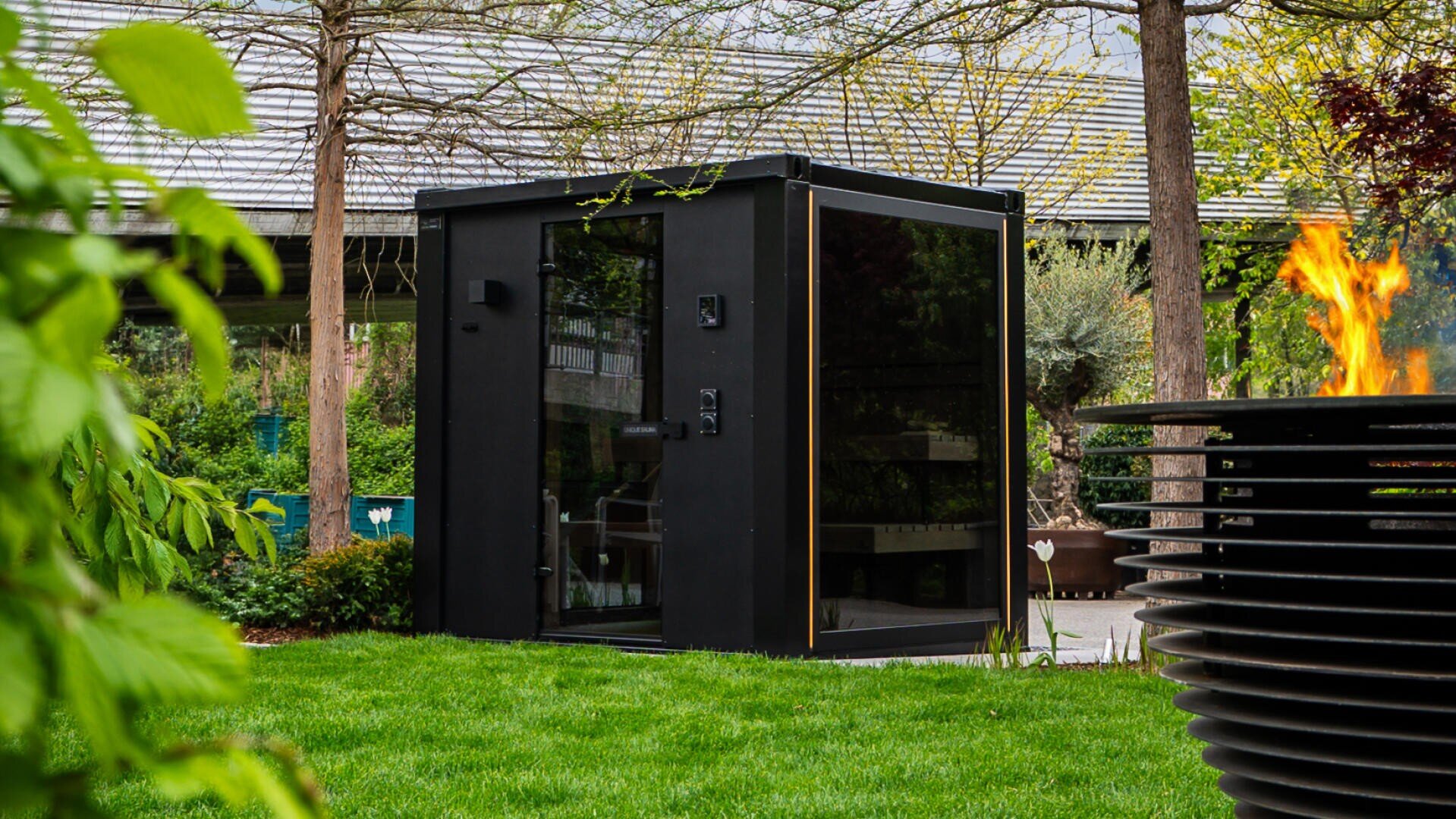 Ein modernes schwarzes Gartenbüro mit großen Glasfenstern steht auf einem grünen Rasen, umgeben von Bäumen und Pflanzen. Im Vordergrund befindet sich eine Feuerstelle mit sichtbaren Flammen.