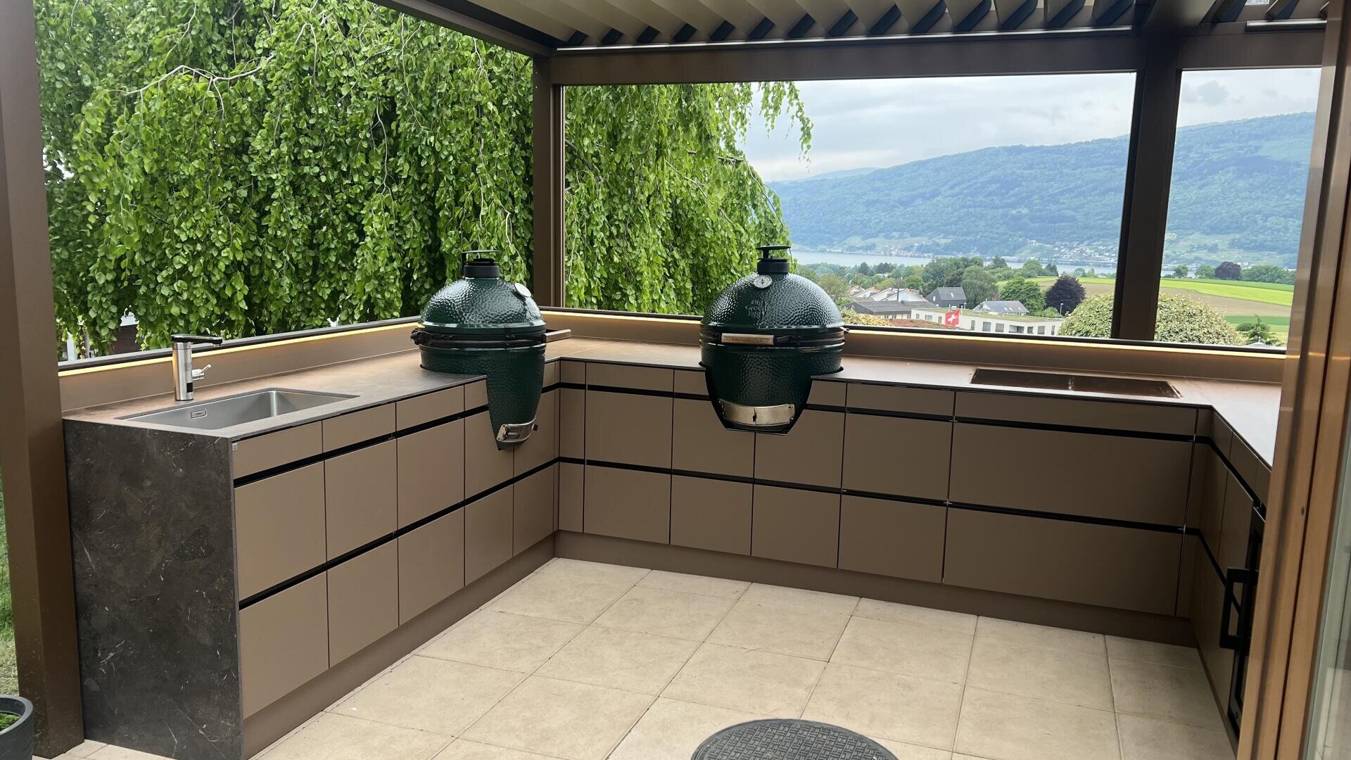Moderne Außenküche mit braunen Schränken, einem eingebauten Spülbecken und zwei großen grünen Keramikgrills. Der Bereich ist überdacht und bietet einen malerischen Blick auf grüne Hügel, Bäume und ein Tal im Hintergrund.