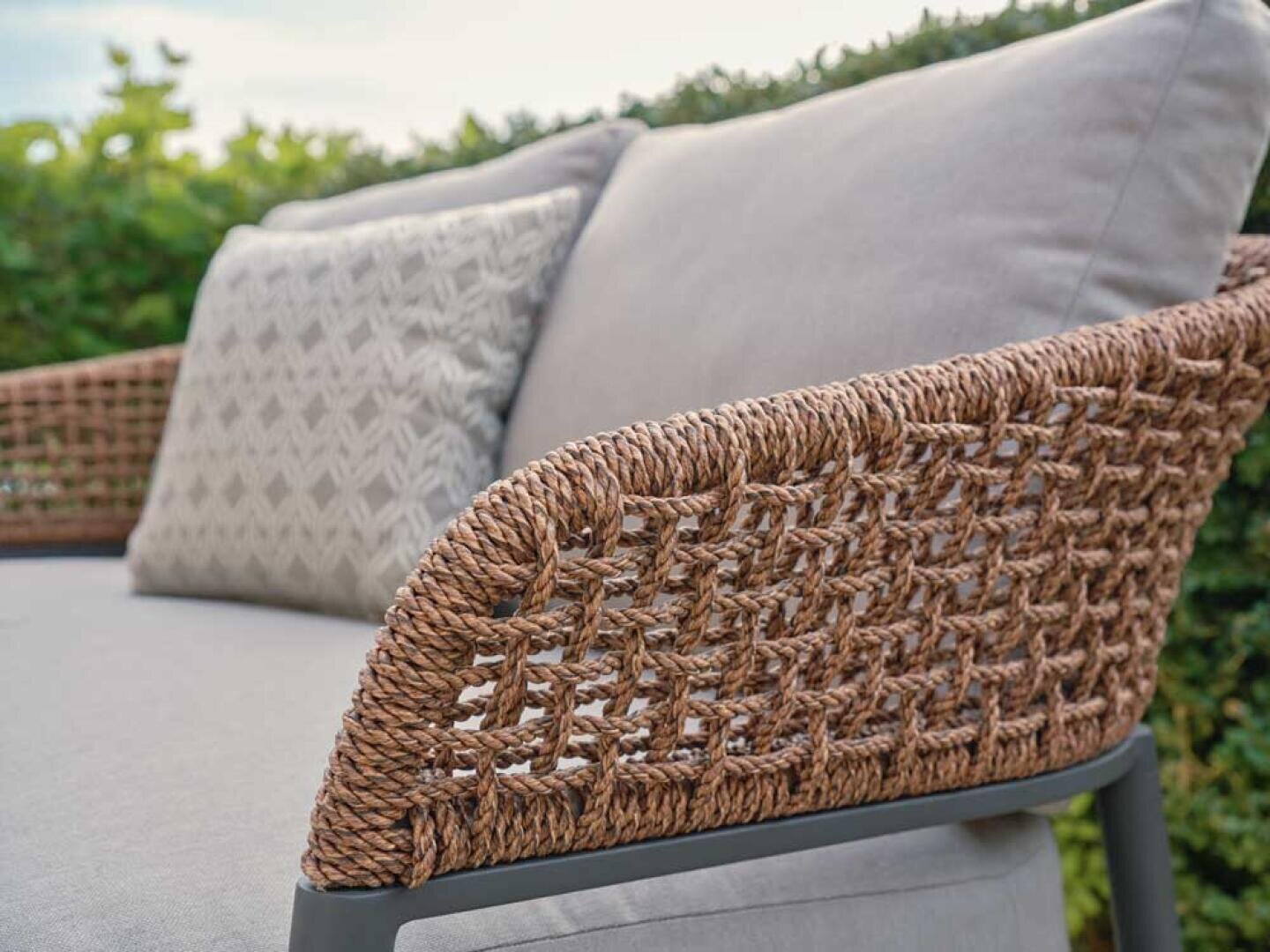 Nahaufnahme eines Korbsofas für den Außenbereich mit grauen Kissen und einem dekorativen gemusterten Kissen vor einem grünen Hintergrund.