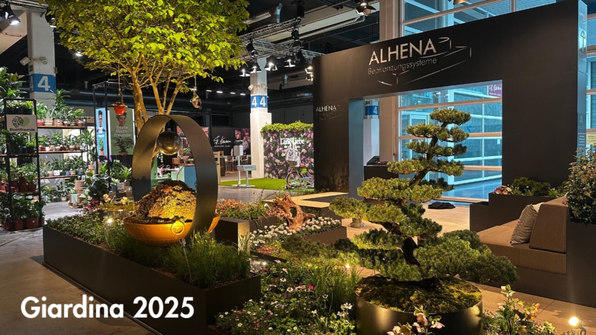Eine moderne Indoor-Gartenausstellung in der Giardina 2025 zeigt Bonsai-Bäume, Topfpflanzen und eine dekorative Kugel, wobei die Marke ALHENA auf einer schwarzen Wand zu sehen ist. Große Fenster und nummerierte Säulen sind zu sehen.