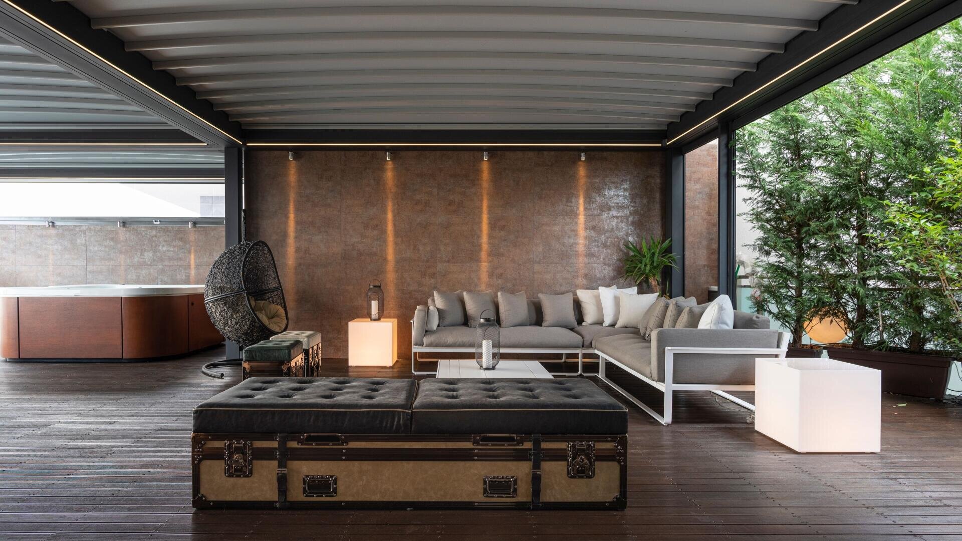 Moderne Outdoor-Lounge mit einem grauen Sektionssofa, dekorativen Kissen, Ottomane, beleuchteten Beistelltischen, einem hängenden Eiersessel, Topfpflanzen und einem Holzboden unter einer überdachten Terrasse mit Bepflanzung auf einer Seite.