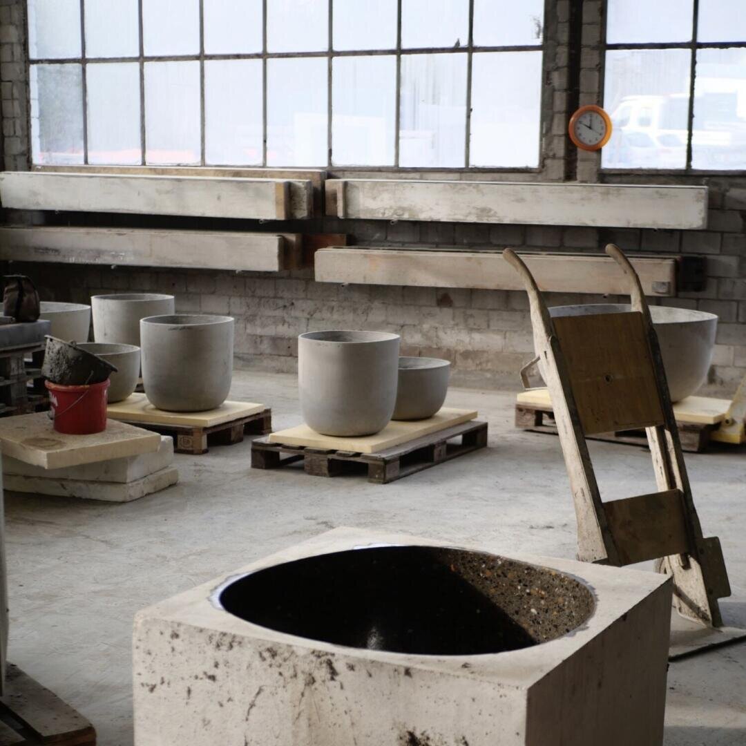 Eine Werkstatt mit großen Betonpflanzgefäßen auf Holzpaletten, einer Schubkarre und Betonblöcken. Hohe Fenster lassen natürliches Licht herein, und an der Rückwand sind Holzbalken angebracht. Ein roter Eimer steht neben den Pflanzgefäßen.