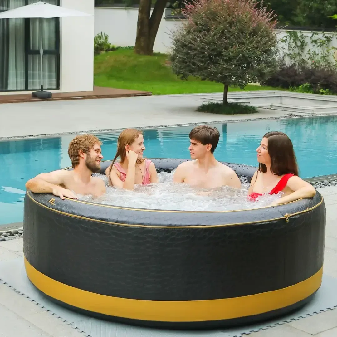 Four people sit and smile in a round, black and gold inflatable hot tub beside a swimming pool in a backyard, with greenery and a modern white building in the background.