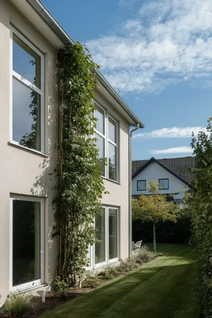 A modern two-storey house with large windows and a vertical green vine growing along the wall, framed by a neatly trimmed lawn and a small garden, under a partly cloudy blue sky. Another house can be seen in the background.