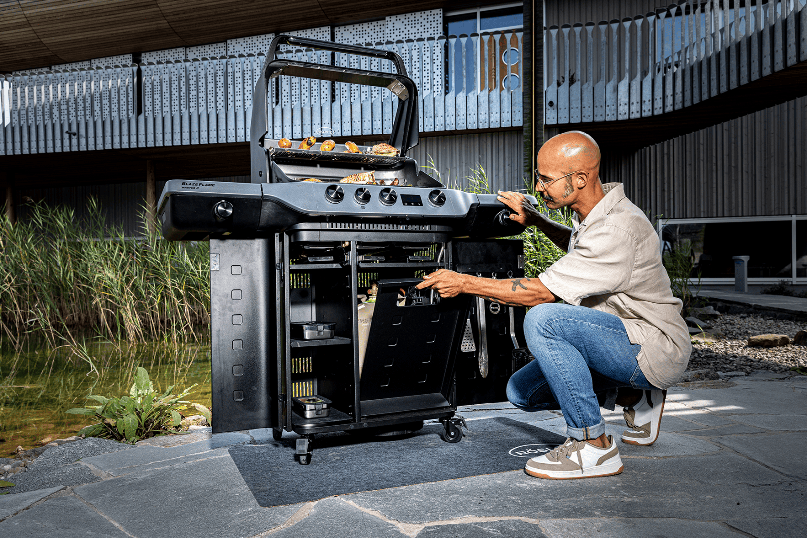 A person kneels next to a black outdoor grill and places a component under the grill. The food is being cooked on the grill. The scene takes place on a stone terrace next to a pond and a modern building.