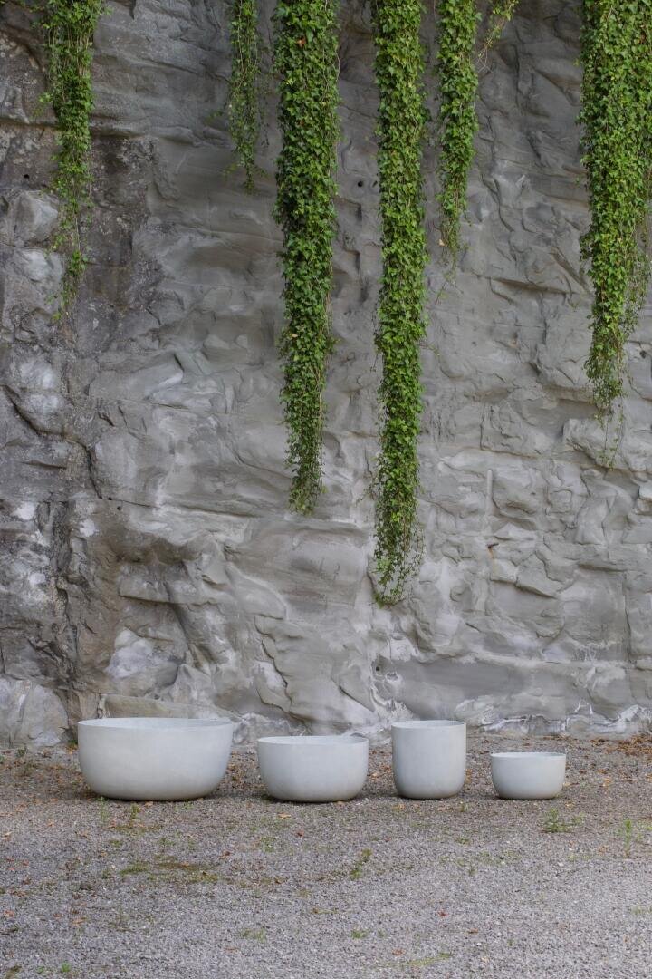 Vier weiße, minimalistische Keramiktöpfe in verschiedenen Größen stehen in einer Reihe auf Kies vor einer grauen Steinmauer, von der lange grüne Ranken herabhängen.