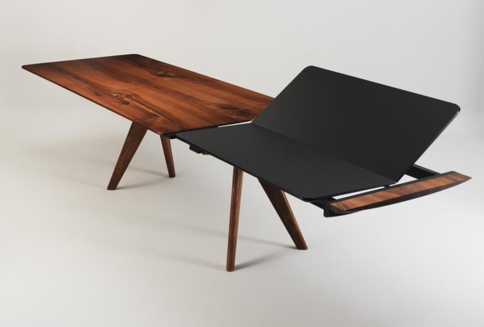 A modern wooden dining table with angled legs and an extended black top that is partially folded out to reveal its folding mechanism against a plain background.