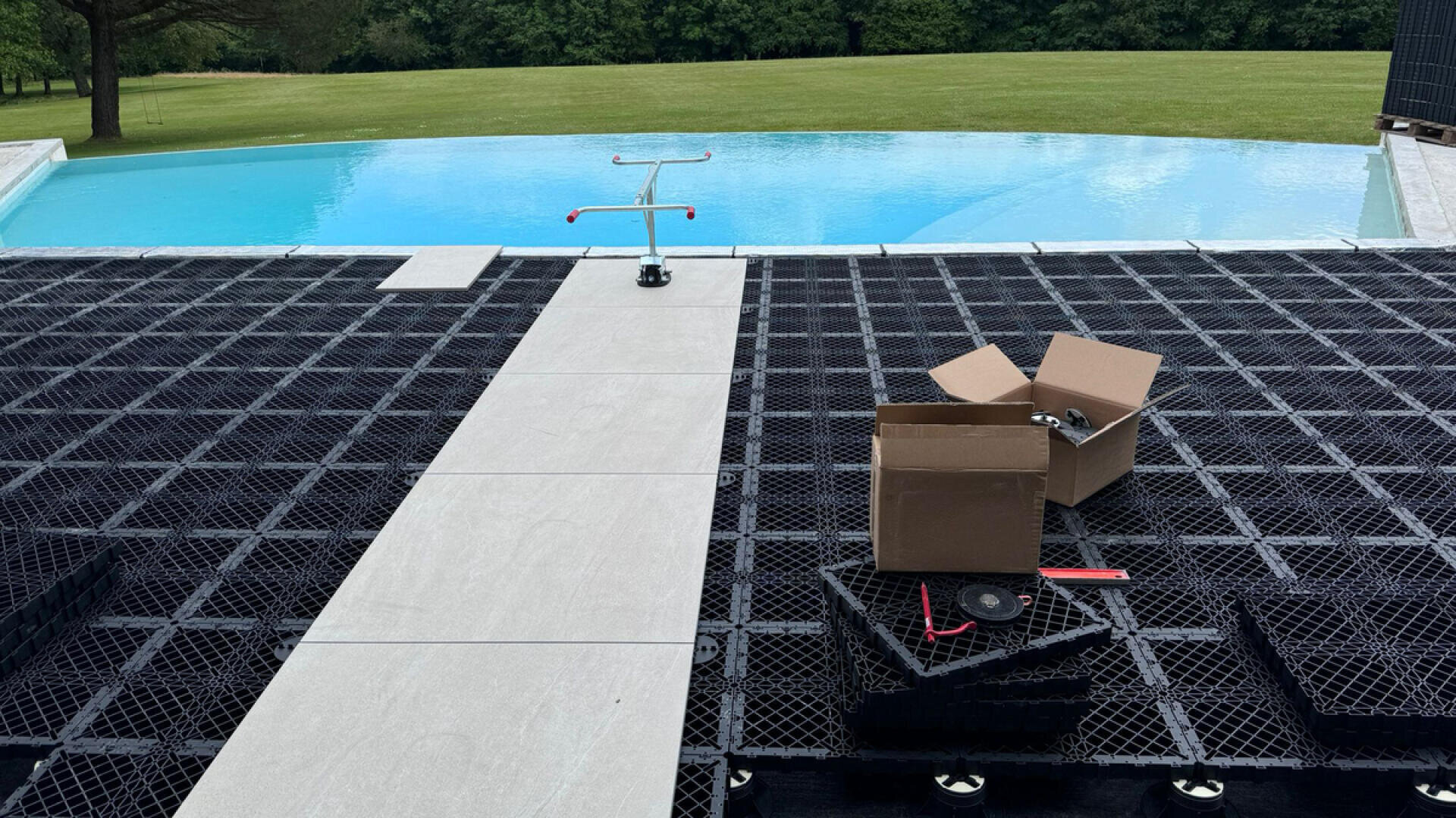 A partially constructed poolside deck with black plastic grid panels, a narrow tile walkway, an open cardboard box with tools and parts, and an infinity pool overlooking a grassy field.