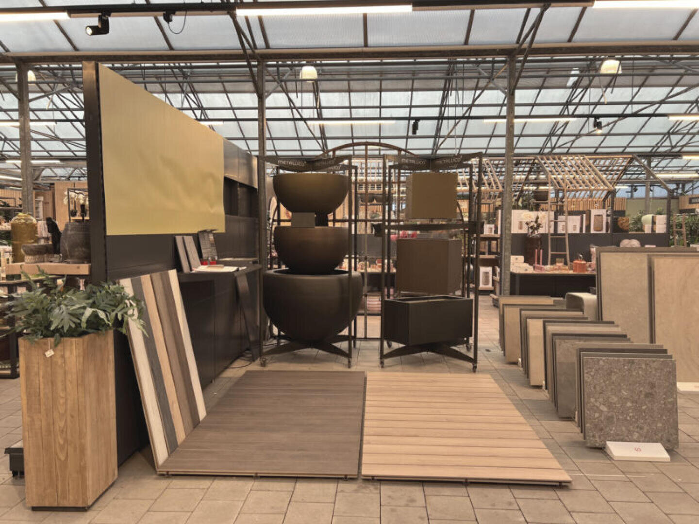 Auslage in einem Baumarkt mit verschiedenen Bodenbelagsmustern, großen gestapelten Pflanzgefäßen, Topfpflanzen und Einrichtungsgegenständen, die auf einem gefliesten Boden unter einem gewächshausähnlichen Dach angeordnet sind.