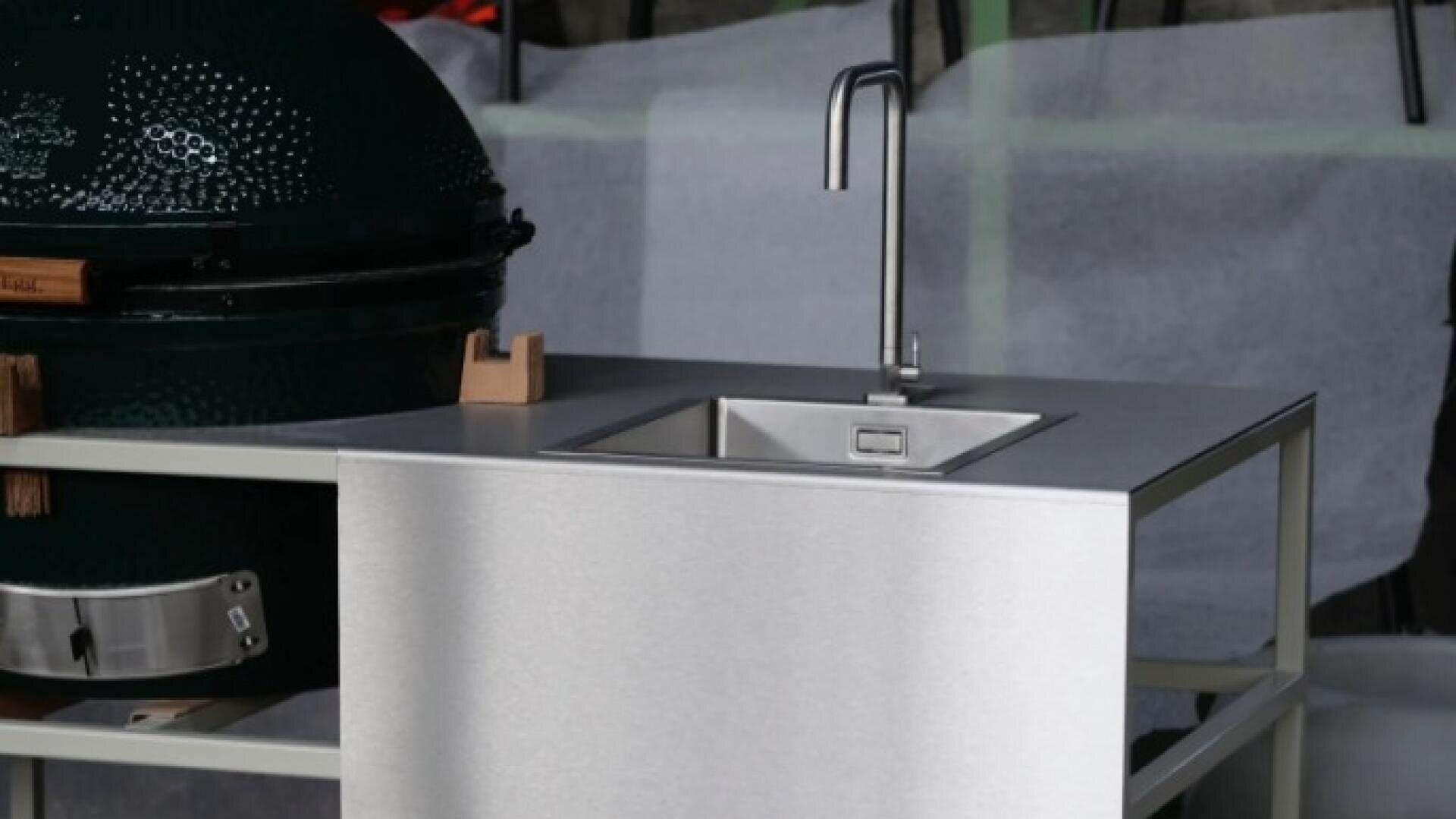 A modern outdoor kitchen setup features a stainless steel sink with a tall faucet next to a green ceramic grill, all set on a metal countertop.