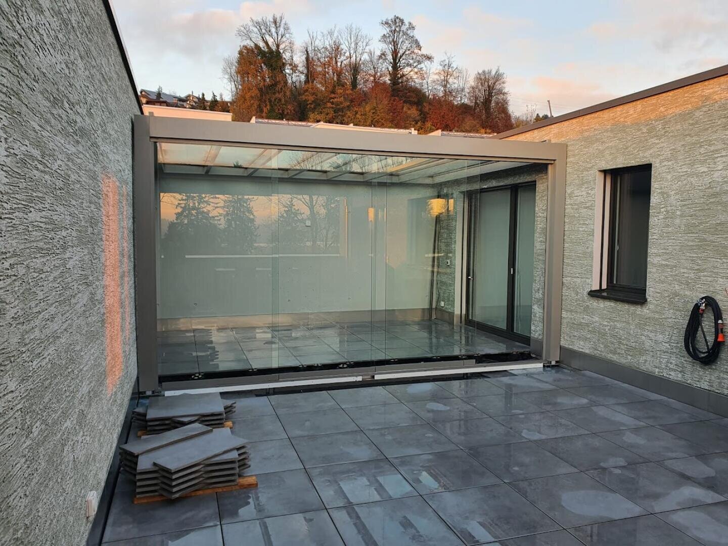 Eine moderne Außenterrasse mit großen grauen Fliesen, einer überdachten Glasabdeckung und gestapelten Fliesen in der Ecke. An der strukturierten Wand ist ein Schlauch angebracht, und im Hintergrund sind Bäume zu sehen.