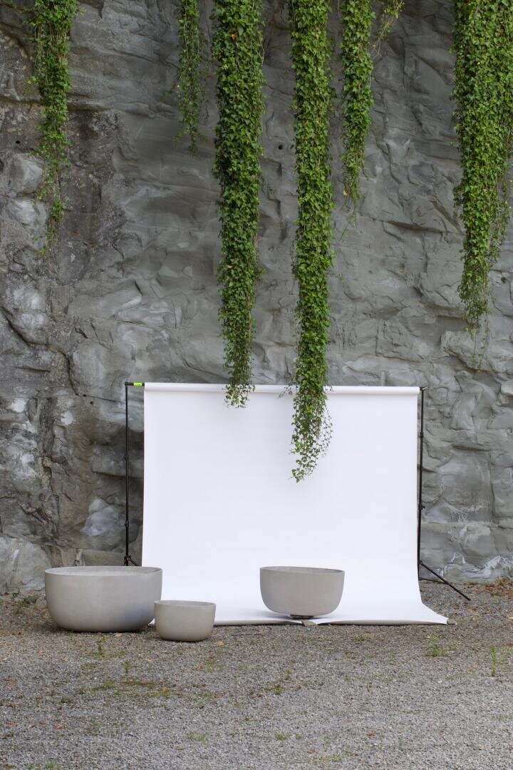 Ein weißer Hintergrund steht vor einer felsigen Wand mit grünen Ranken darüber, während drei große, leere graue Pflanzentöpfe auf Kies vor dem Hintergrund platziert sind.
