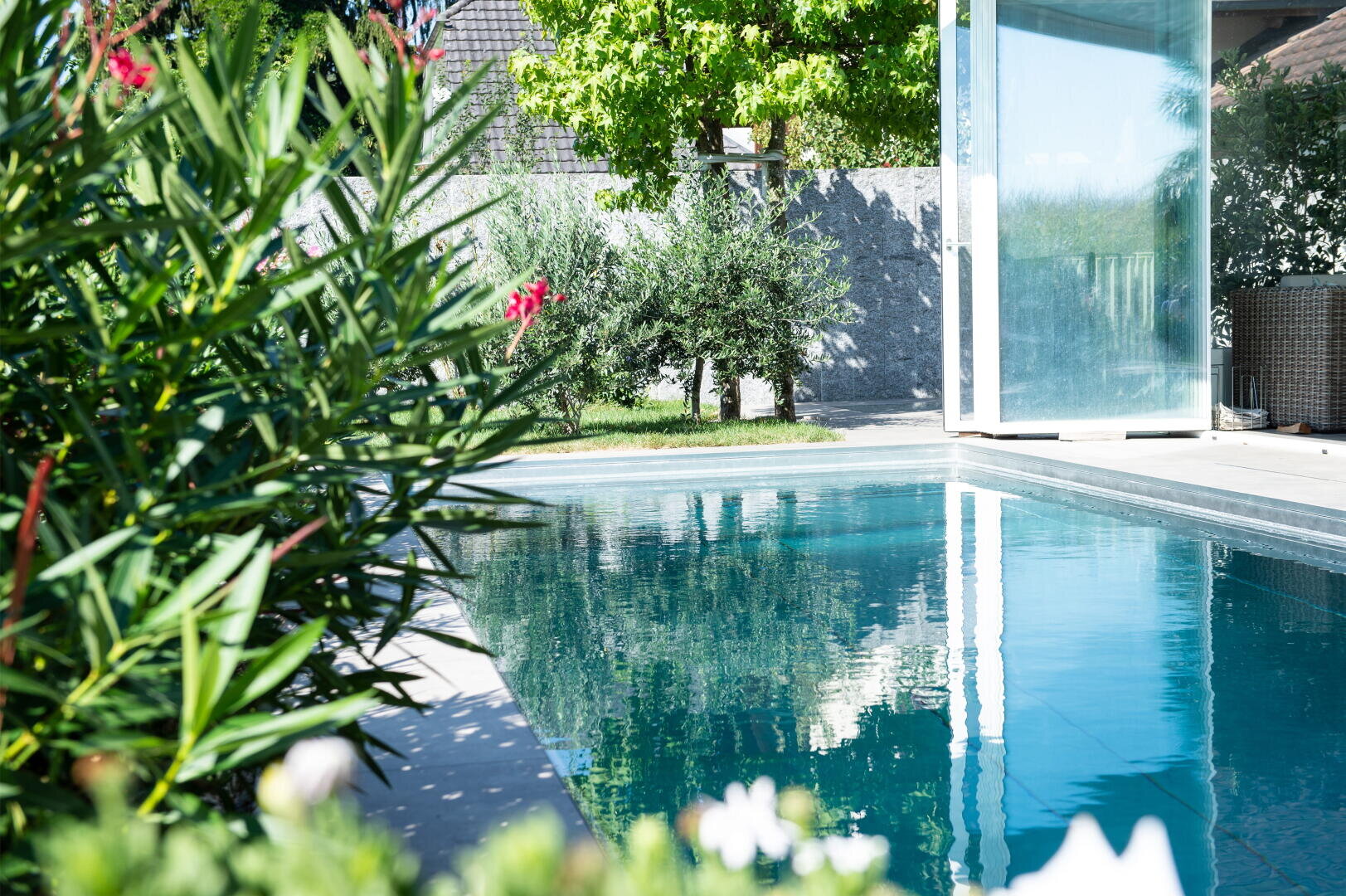 Ein moderner Swimmingpool im Freien neben einem Garten mit grünen Pflanzen und Bäumen. Das Sonnenlicht spiegelt sich im Wasser, und eine Glastür führt auf die Terrasse.