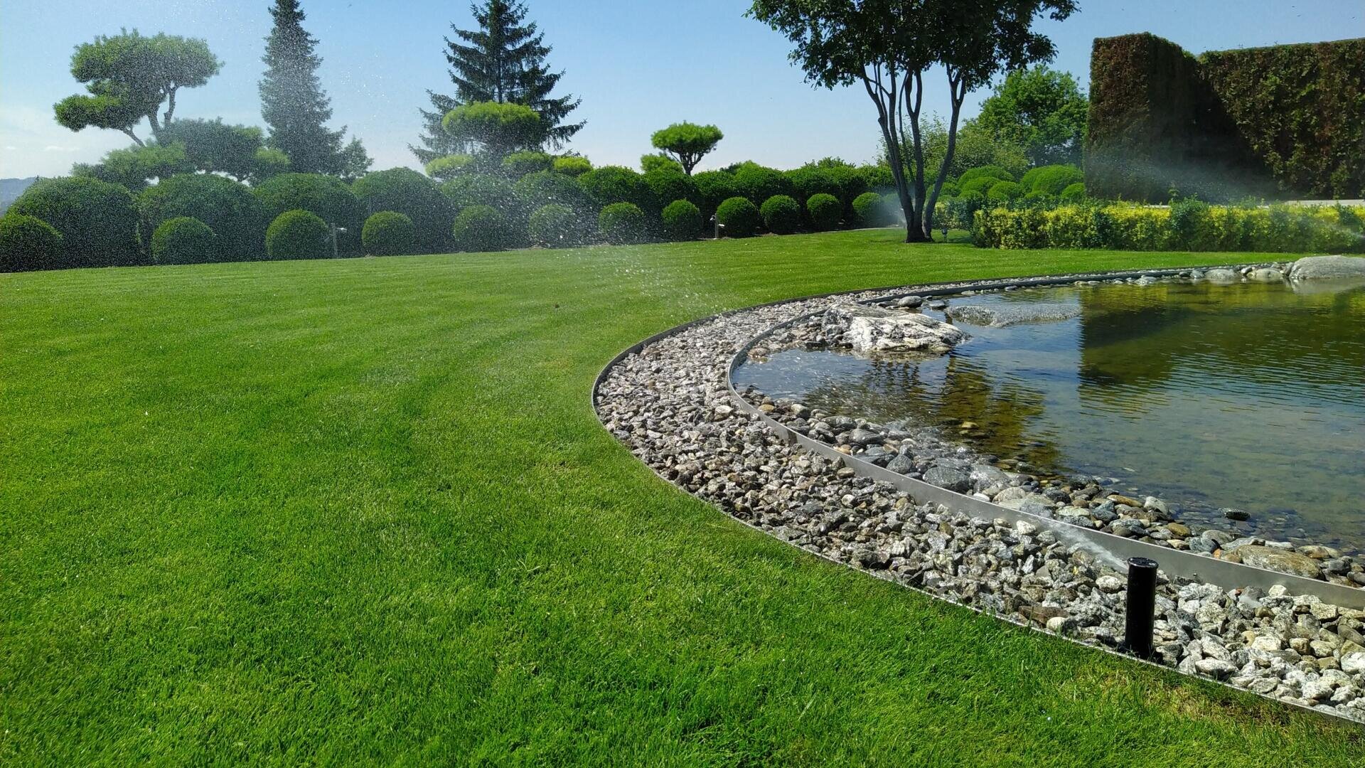 Ein angelegter Garten mit gepflegtem Rasen, einem kleinen, mit Steinen eingefassten Teich, ordentlich gestutzten Büschen und hohen Bäumen im Hintergrund unter einem klaren blauen Himmel. Sprinkleranlagen bewässern den Rasen.