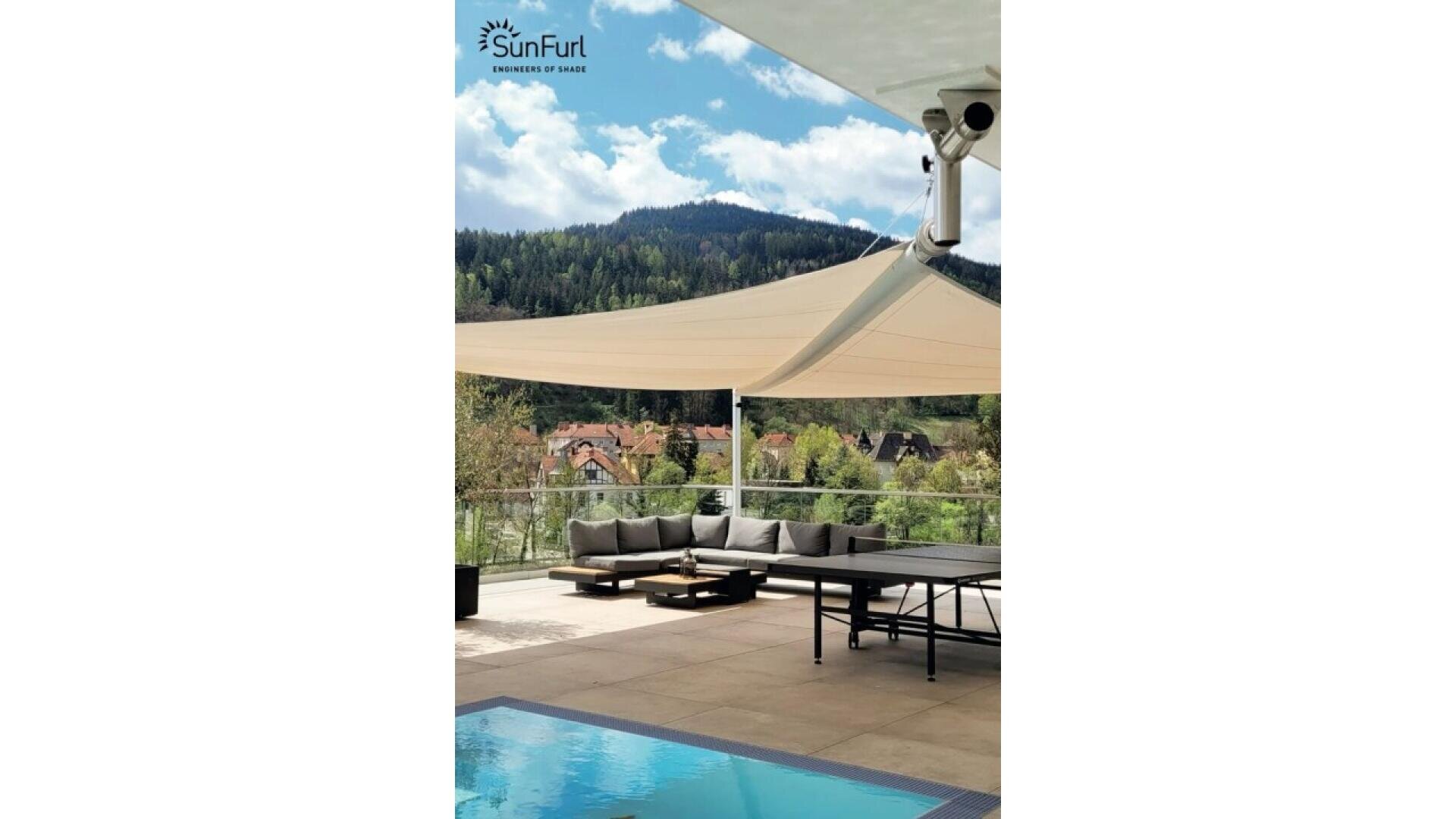A modern patio with a pool, shaded by large sail canopies, features a gray outdoor sofa and ping pong table. Behind, green hills and scattered houses are visible under a partly cloudy sky. SunFurl logo appears at the top left.