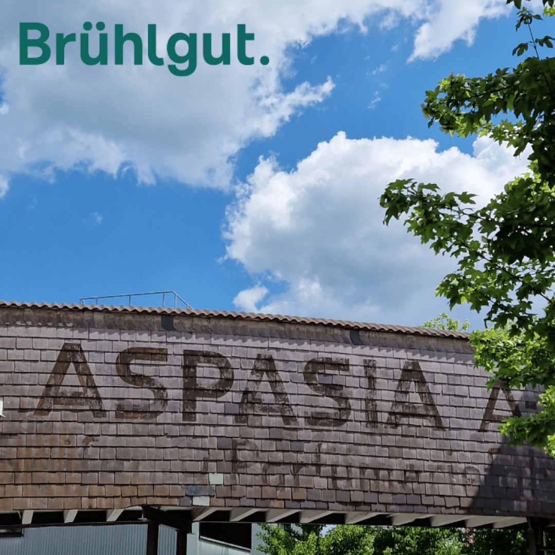 A sunny sky with white clouds over an old building with a tiled roof and a faded ASPASIA sign. Green tree branches frame the right side. Brühlgut. is written in green at the top left.