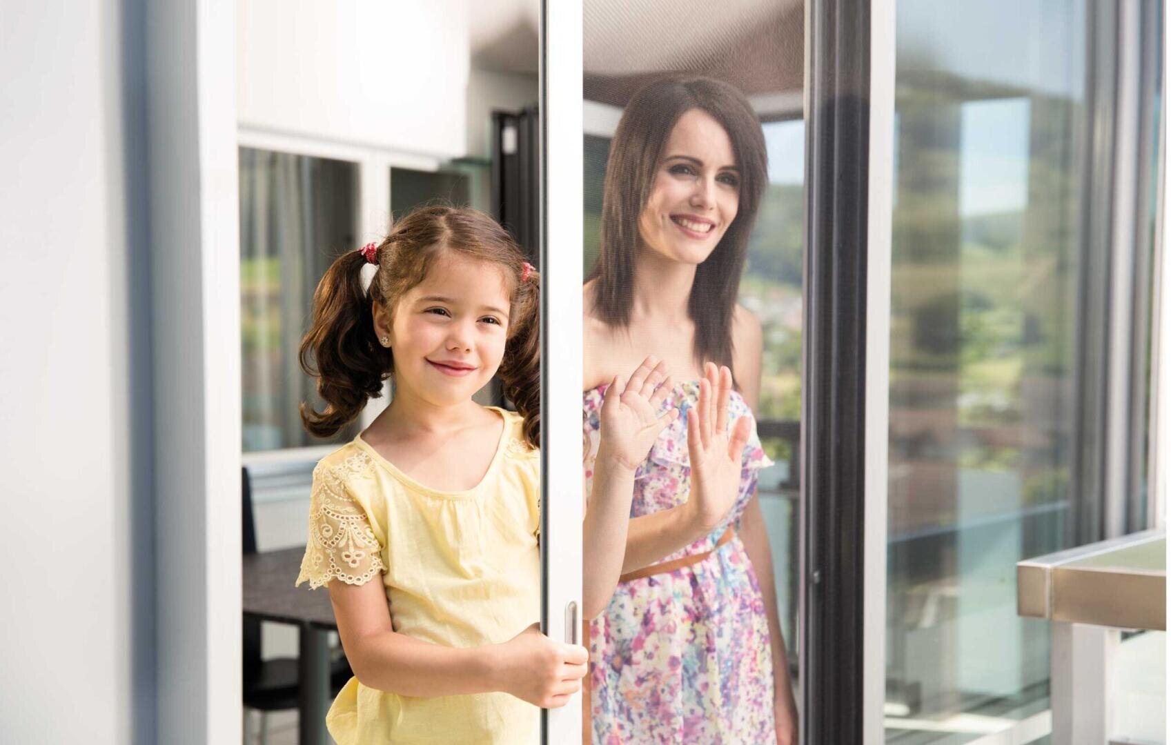 Ein junges Mädchen in einem gelben Hemd steht lächelnd an einer Glasschiebetür, während eine Frau in einem geblümten Kleid lächelt und das Glas von außen berührt.