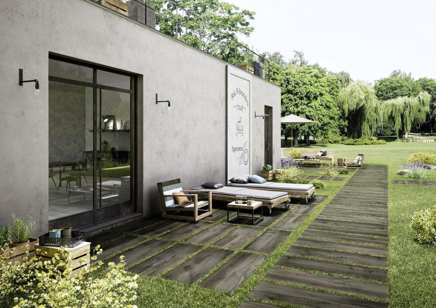 Moderner Innenhof mit Holzmöbeln und Liegestühlen auf einem gefliesten Weg, neben einem Gebäude mit großen Glastüren, mit Blick auf einen üppigen grünen Garten mit Bäumen und Blumen.