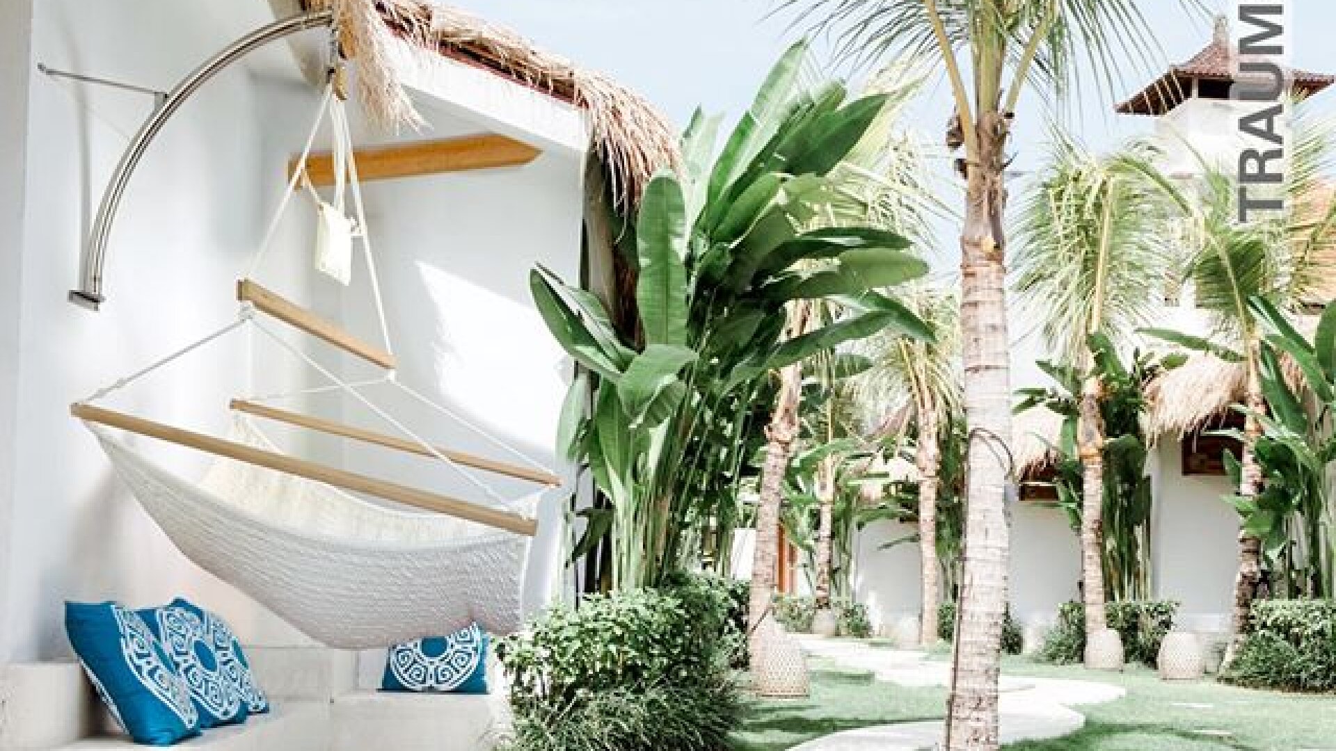 Ein heller, tropischer Innenhof mit Palmen, üppigen grünen Pflanzen und weißen Gebäuden. Eine weiße Hängematte mit blau gemusterten Kissen hängt an einer Wand und schafft eine entspannte, einladende Atmosphäre.