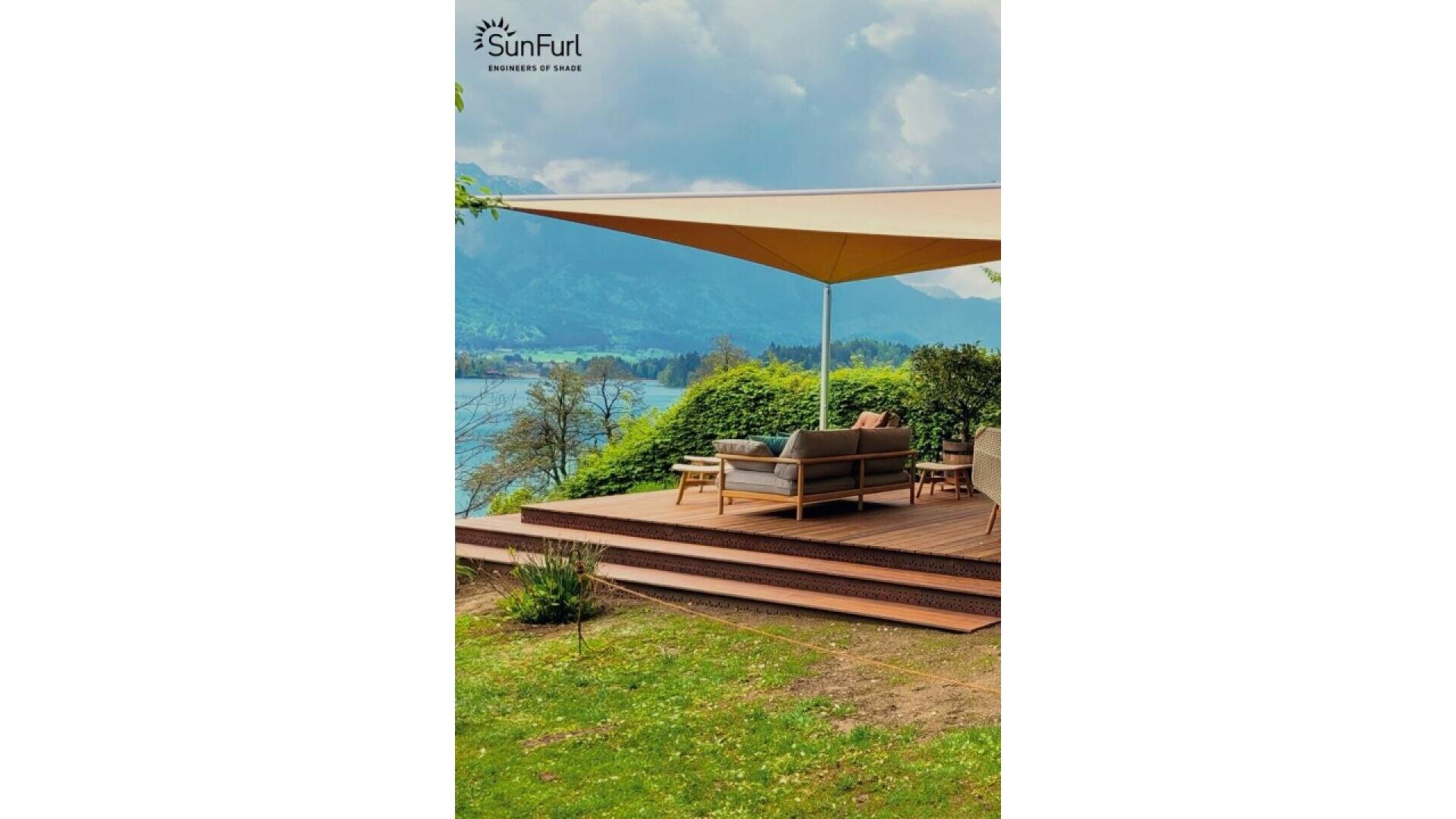 A wooden deck with outdoor seating under a large sunshade overlooks a scenic lake and mountains, surrounded by lush greenery. The SunFurl logo appears at the top of the image.