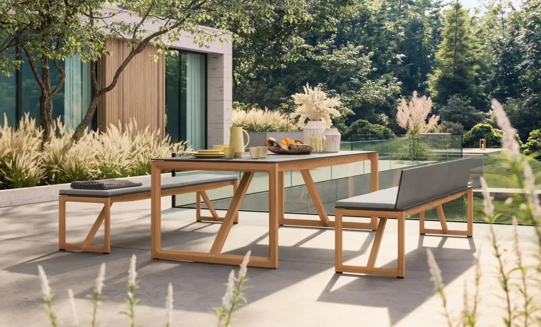 Modern outdoor patio with a wooden table set, including benches and a stool, arranged on a concrete terrace. Table is set with breakfast items and a vase of flowers, surrounded by lush greenery and tall grasses.